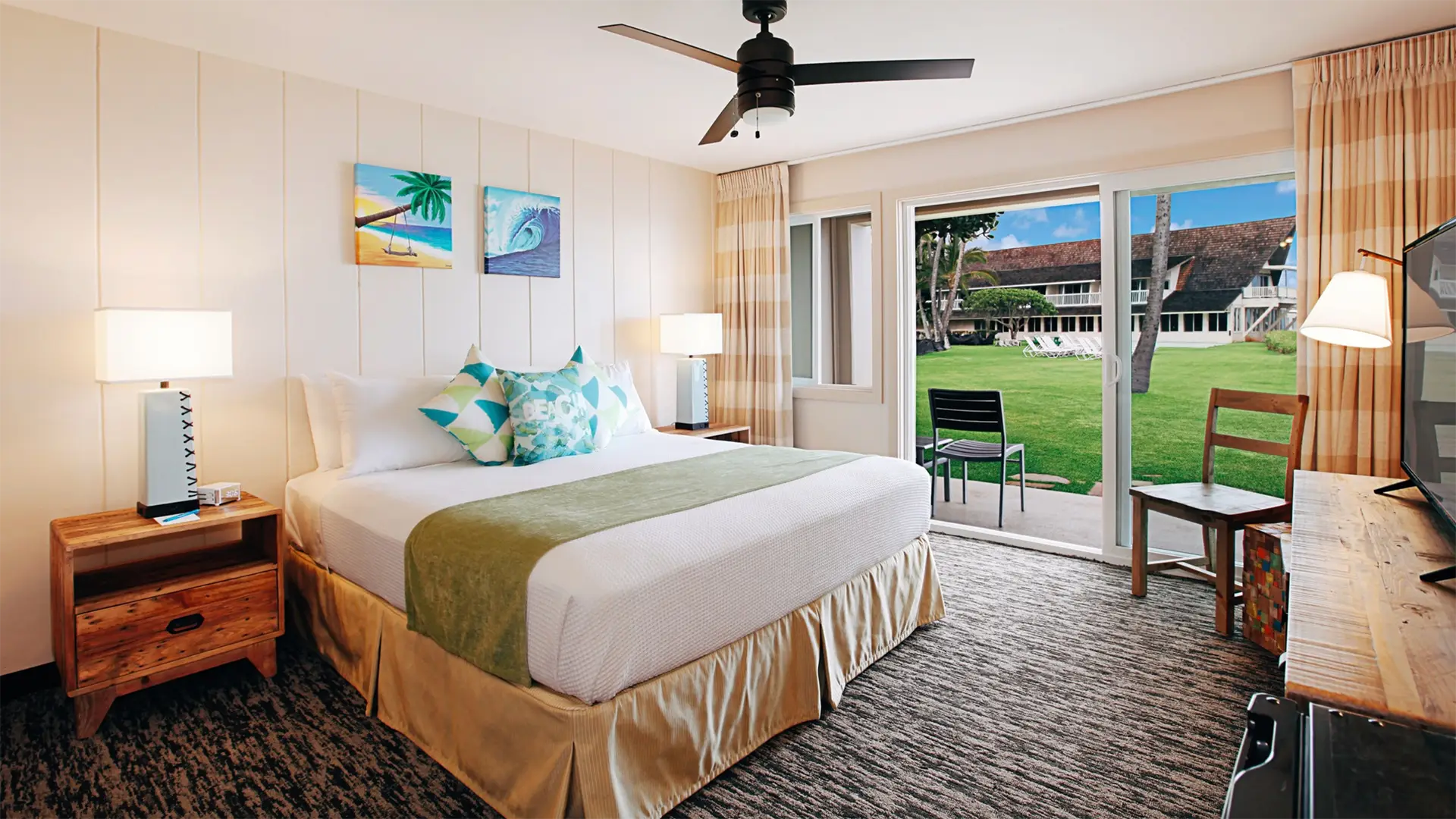 A well-lit hotel room with a large bed adorned with green and white pillows, wooden nightstands with lamps, a ceiling fan, and a painting on the wall. There is a sliding glass door that opens to a grassy area with tables and chairs outside. Experience one of the best value hotels Kauai has to offer.