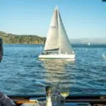 A couple is seated at a dining table by a window, gazing at a white sailboat gliding on the calm blue waters of Tiburon. The lush, green coastline is visible in the background under a clear blue sky.