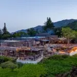 A scenic outdoor restaurant nestled in a lush, green mountainous area. The restaurant features wooden structures, string lights, and numerous tables. Surrounding vegetation and evening lighting create a cozy and inviting atmosphere, reminiscent of the best luxury hotels on the Monterey Peninsula.