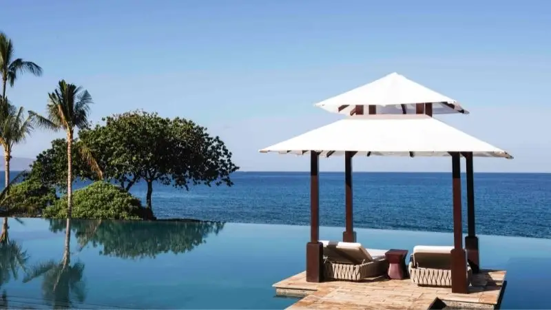 An infinity pool overlooking a serene ocean. The polished poolside features a shaded cabana with white fabric roofing, wooden posts, and two cushioned lounge chairs. Palm trees and lush greenery stand on the left side, under a clear blue sky—truly one of the best family hotels Maui has to offer.