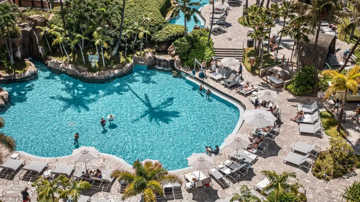 A lively aerial view of a tropical resort swimming pool area at one of the best oceanfront hotels in Maui. The large pool is surrounded by palm trees, sun loungers, and umbrellas. Guests are swimming, lounging, and enjoying the sunny day. Pathways and lush greenery add to the tranquil atmosphere.