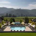 A luxurious outdoor pool area at a resort is surrounded by sun loungers and umbrellas, reminiscent of the elegance found at Napa hotels. Two parallel buildings flank the pool, with a scenic view of lush greenery and rolling hills under a clear sky in the backdrop.