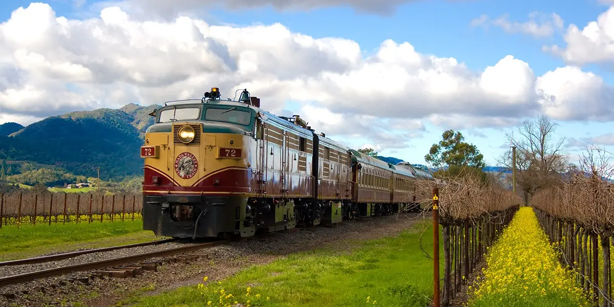 A vintage train with red and yellow locomotives moves through a scenic countryside with mountains in the background. The tracks are surrounded by vineyards and blooming yellow flowers, making it an idyllic way to explore wine country under a partly cloudy sky.