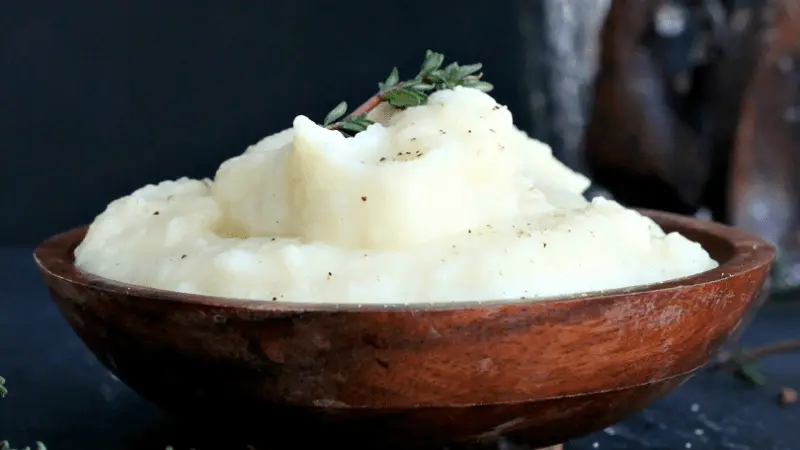 A wooden bowl filled with creamy mashed potatoes, a staple of our winter menu, is garnished with a sprig of thyme and set against a dark background. The mashed potatoes have a smooth texture and are lightly speckled with black pepper.