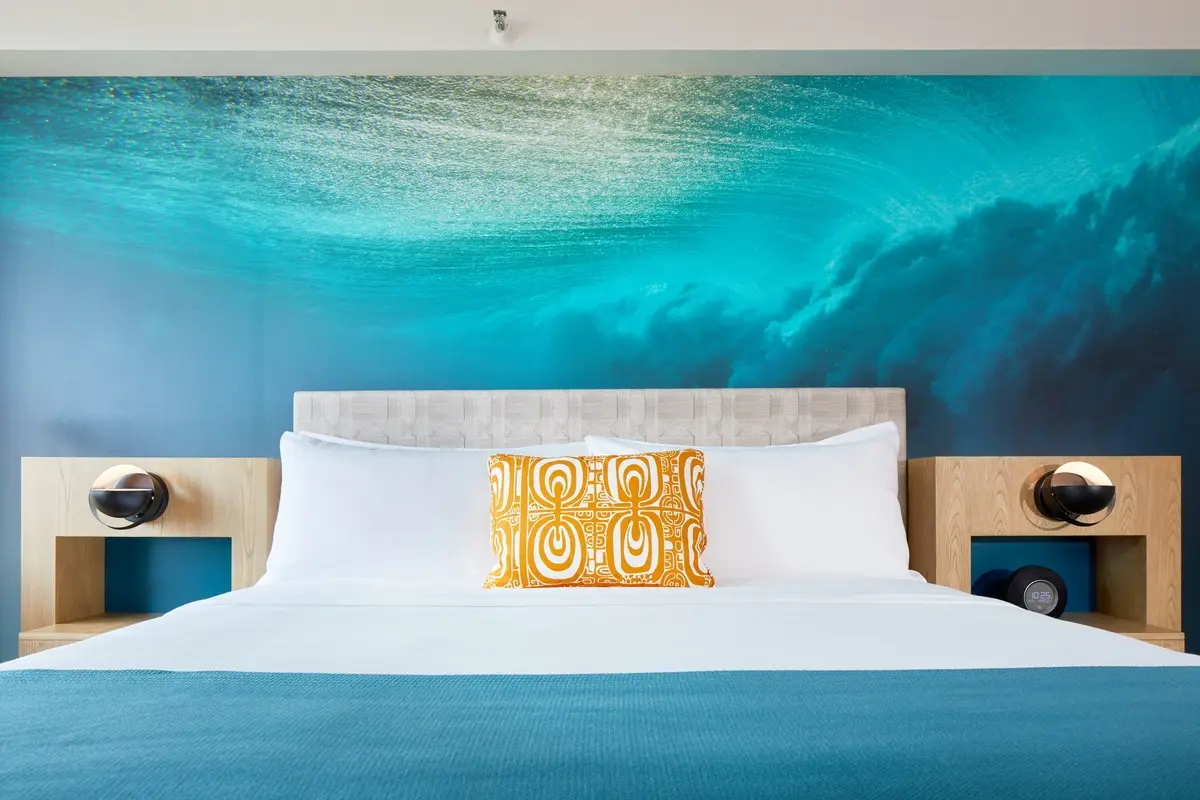 A stylish bedroom in this Hawaii home features a bed with white linens and a yellow patterned pillow. The headboard is set against a mural of an underwater scene, creating a tranquil atmosphere. Modern bedside tables are adorned with spherical lamps and small decorative items, reminiscent of Waikiki Beachcomber style.