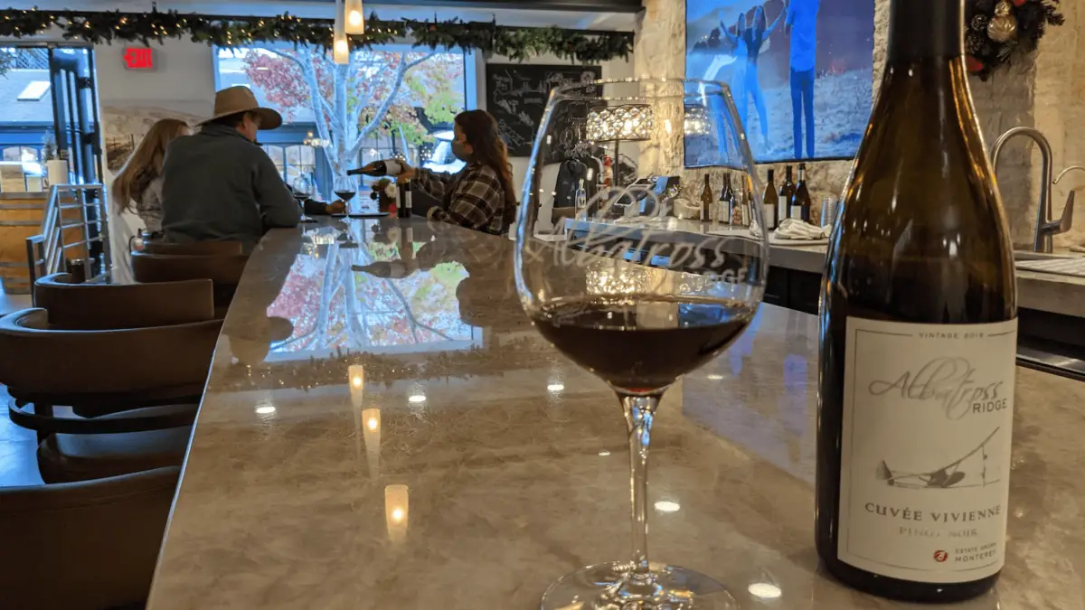 A wine glass filled with red wine and an open bottle labeled "Albatross Ridge Cuvée Vivienne" sit on a polished bar counter. In the background, people are seated and engaging in conversation at the warmly lit Carmel Wine Tasting Rooms.