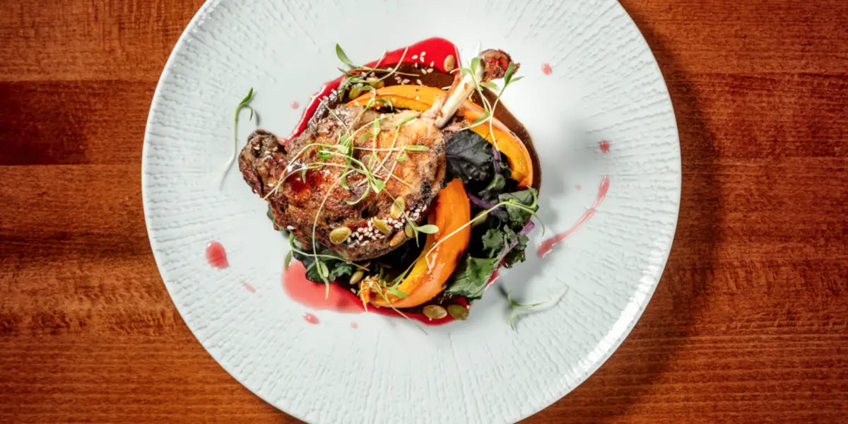 A beautifully plated dish featuring succulent, roasted meat garnished with microgreens, presented with vibrant orange and yellow roasted vegetables, all arranged on a white plate with a splash of red sauce. This culinary gem hails from one of those under-the-radar Bay Area towns.