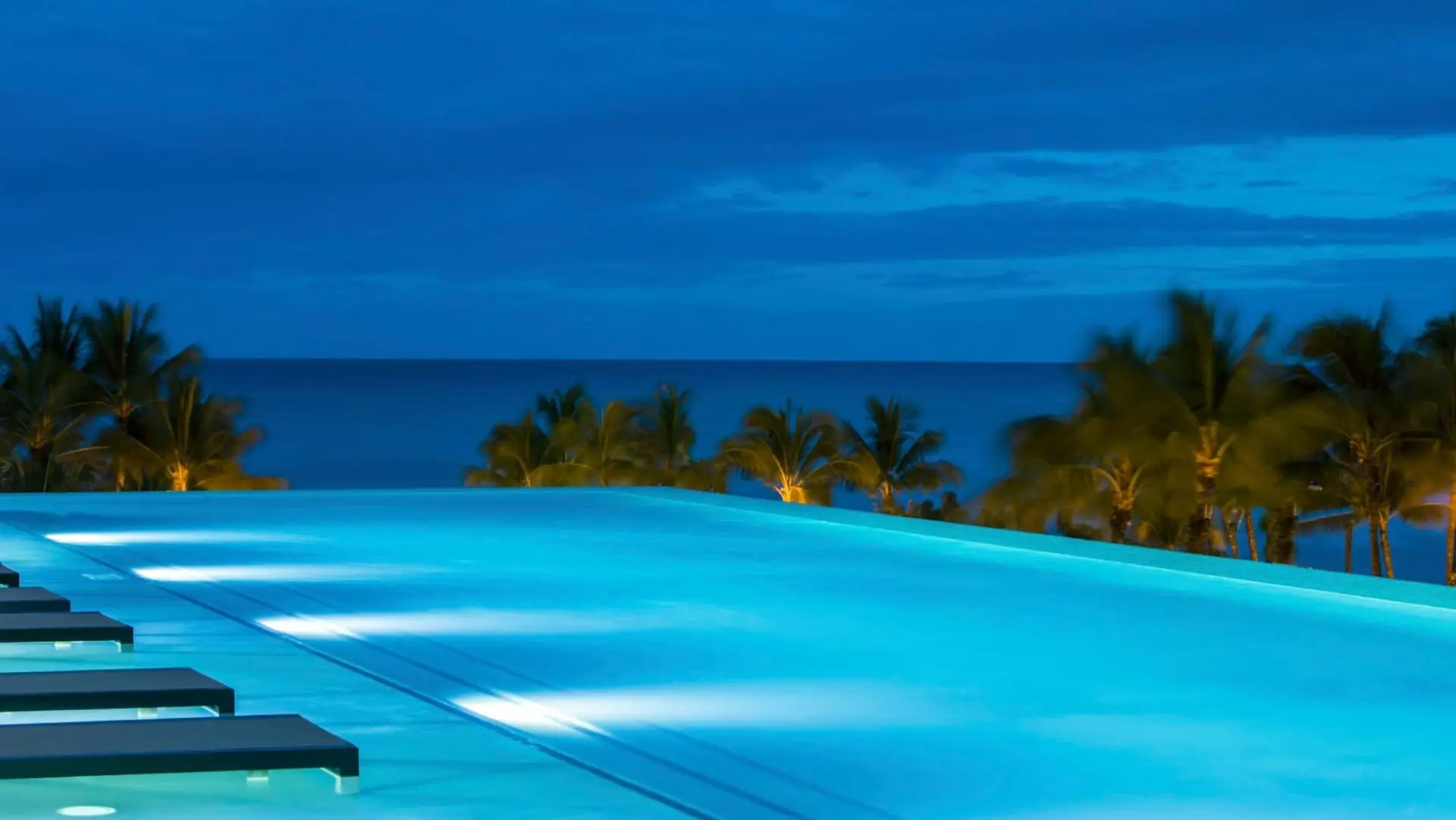 An infinity pool at night appears to blend seamlessly with the blue horizon of the ocean, framed by the silhouettes of palm trees. Rated among the best hotel pools in Oahu, it is illuminated and casts a serene glow over the water, creating a tranquil and picturesque tropical setting.