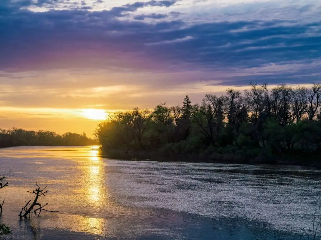 A serene river at sunset, with the sun partially hidden behind silhouettes of trees on the riverbank. The sky displays shades of purple, blue, and orange, reflecting on the calm water—creating a tranquil and picturesque scene straight out of *The Ultimate Guide to Sacramento*.