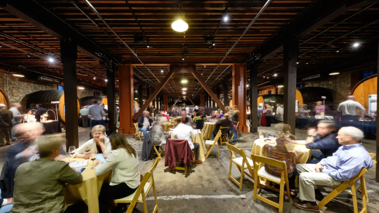 A vibrant, busy indoor event captures people dining and mingling at tables set in a spacious, rustic venue. Long tables are aligned under warm lighting, with wooden beams and large wine barrels visible in the background, creating an inviting, lively atmosphere. One of the best things to do in wine country this April.