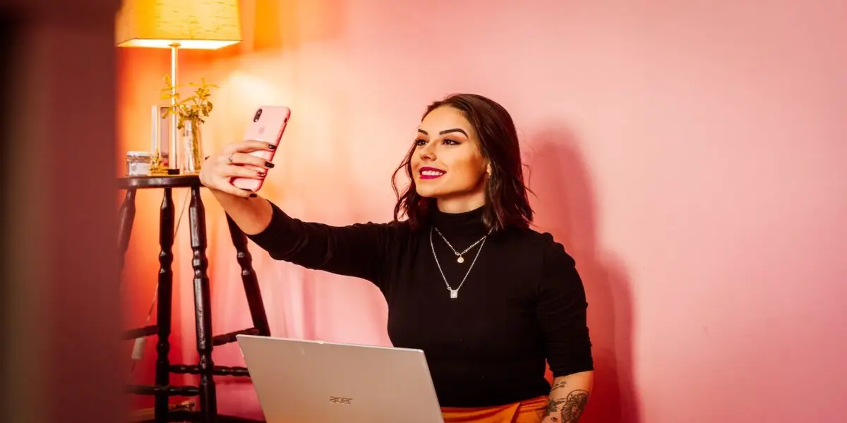 A woman with shoulder-length hair sits in front of a laptop, taking a selfie with her phone on Gen Z’s new favorite app. She is wearing a black turtleneck and has a tattoo on her arm. The background features a pink wall and a lamp on a side table. She smiles and looks at the phone.