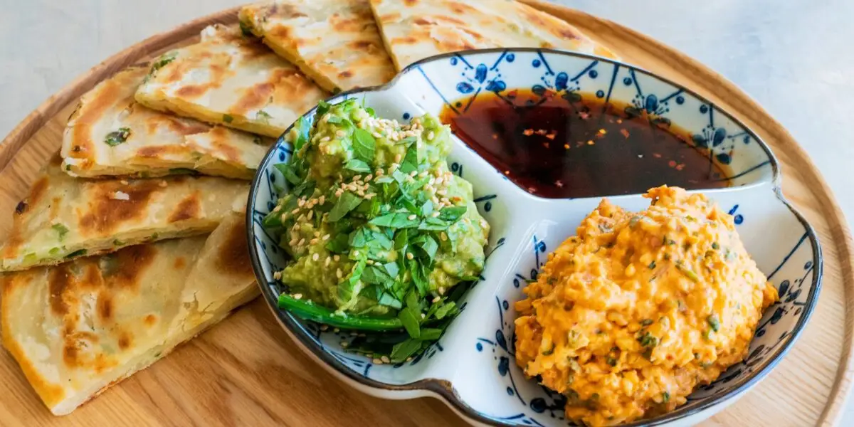 A wooden plate contains sliced scallion pancakes arranged around a divided dish, which holds three compartments of dipping sauces: soy-based sauce, avocado salad with sesame seeds, and a spicy mayo mixture. Perfect for the best dinner in Wine Country, the colorful presentation enhances the appetizing look.
