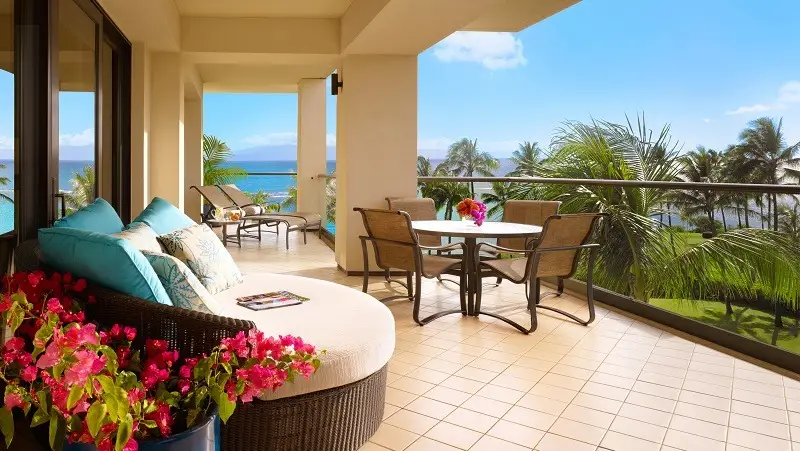 A spacious balcony with beige tiles features a round daybed adorned with blue cushions and a small table with four chairs. Pink flowers decorate the space. The view showcases palm trees and a blue ocean under a clear sky, embodying the charm of the best romantic hotels Maui has to offer.