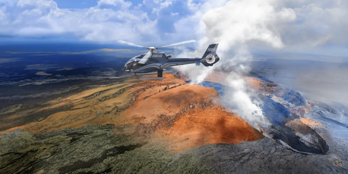 A helicopter flies over an active volcanic landscape with smoke rising from the volcano. The terrain below is a mix of black, brown, and red hues, indicating lava flow and ash. Experience the best helicopter tour on the Big Island as you marvel at this breathtaking sight. The sky is partly cloudy with patches of blue visible.