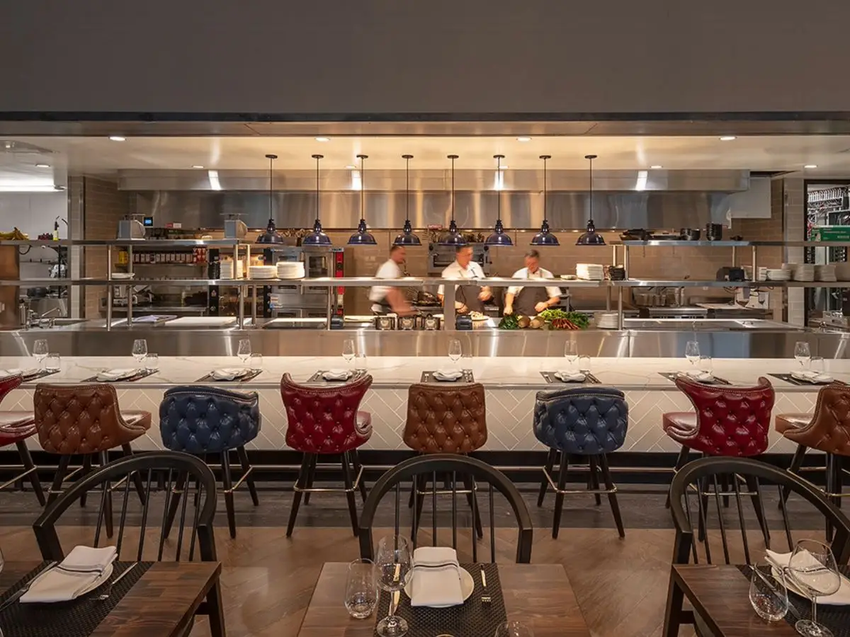 A modern open-kitchen restaurant with leather bar stools in red, blue, and brown arranged in front of the counter. Three chefs are actively working in the background, and tables are set with plates, cutlery, and glasses in the dining area—truly offering the Best Dinner in Sacramento.