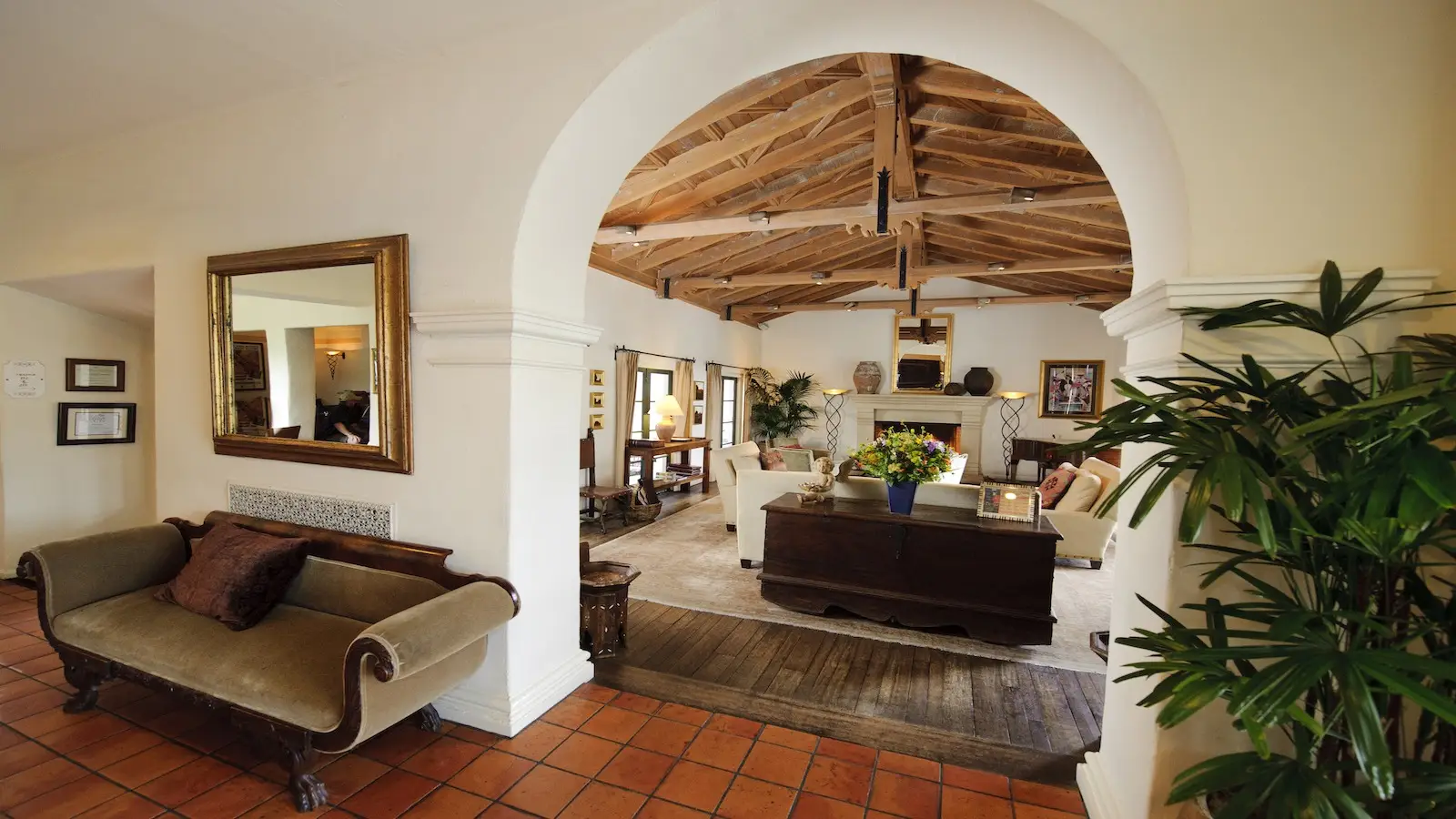 A cozy living room with rustic wooden ceiling beams and white walls evokes the charm of Central California missions. An arched doorway leads to a seating area with sofas, a coffee table, and a decorative fireplace. Potted plants and a large mirror add to the warm, inviting atmosphere.