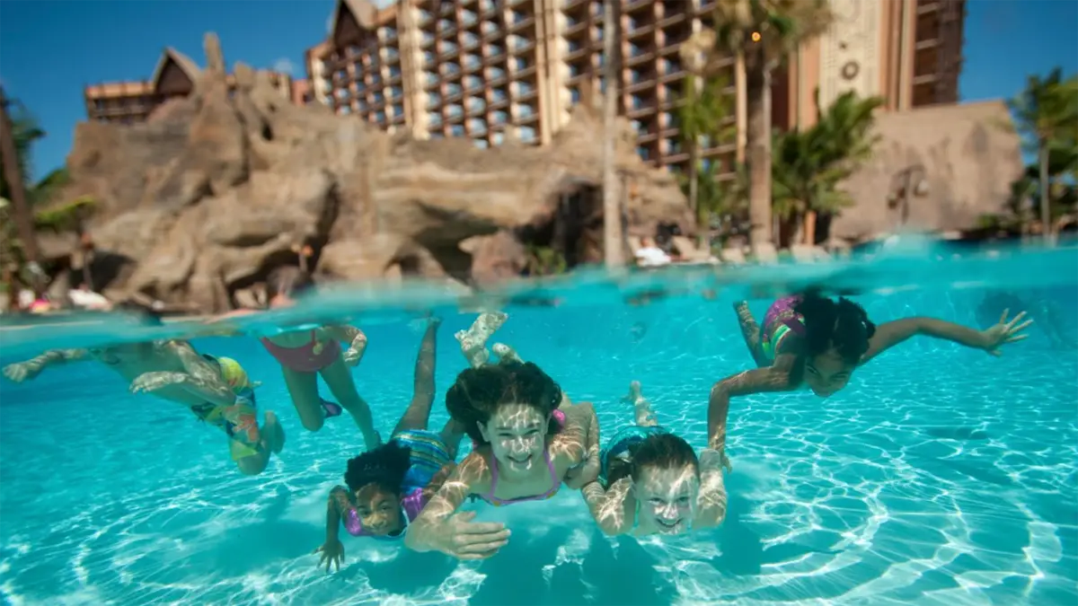 Underwater view of several children swimming in a pool at one of the best family hotels Oahu offers. The background shows a large resort building with numerous windows and a rocky, water slide structure. The children appear to be enjoying their swim with arms and legs splayed out.