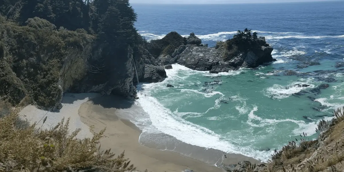 A secluded beach cove in Big Sur with clear turquoise water and white waves gently washing onto the sandy shore. Tall cliffs surround the cove, and lush greenery and trees are perched atop the cliffs. The sky is clear, revealing the vast ocean in the background—truly an ultimate guide to Big Sur's beauty.