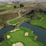 Aerial view of a scenic golf course featuring lush green fairways, sand bunkers, and serene water hazards. The course, one of the best golf courses in the greater Bay Area, is surrounded by vineyards and rolling hills, creating a picturesque landscape under a clear sky. Paths wind through the fields and course.