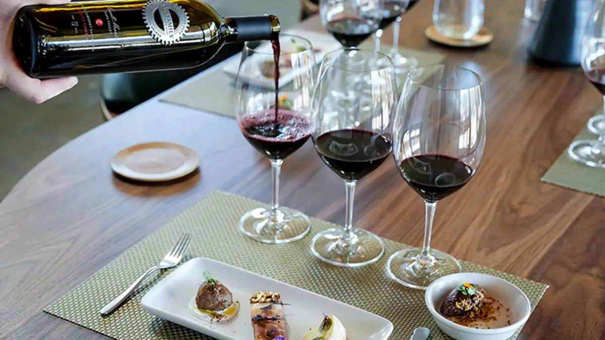 A table setting with three glasses of red wine, a bottle pouring wine into one glass, and a plate with gourmet appetizers. The wooden table has place mats and cutlery set around it, reminiscent of the elegance found in Napa Valley tasting rooms.