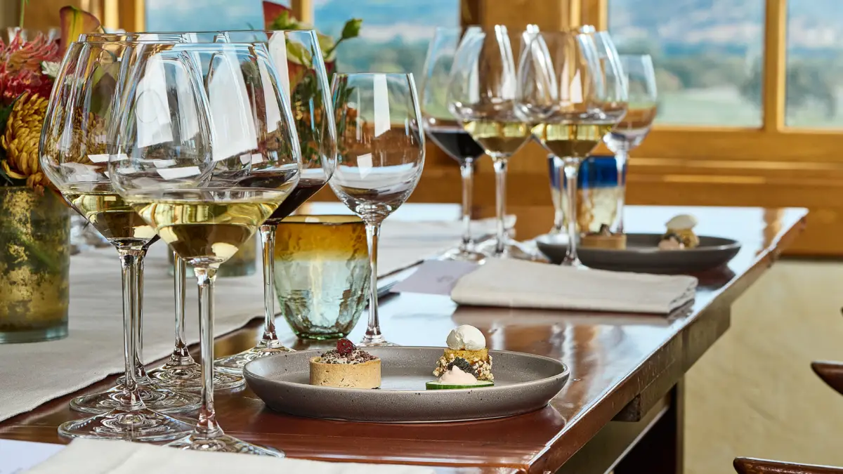 A wooden table set for a meal, featuring multiple wine glasses, small plates of appetizers, candles, and floral arrangements reminiscent of Napa Valley tasting rooms. Sunlight streams in through a window with a scenic view of greenery in the background. White napkins are neatly placed beside the plates.