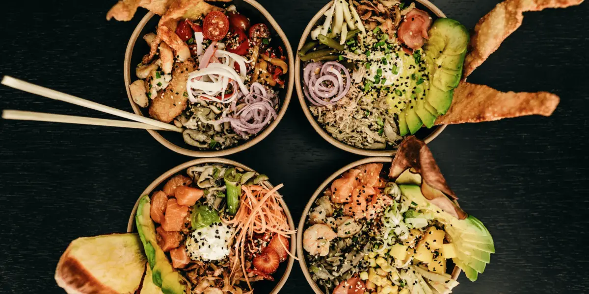Four bowls of colorful and diverse poke bowls are shown from a top view, each filled with a variety of fresh ingredients such as avocado, sliced vegetables, seasoned meats, and rice, garnished with black sesame seeds. Chopsticks rest next to one of the bowls—truly an entry from a Hawaiian Food Glossary come to life.