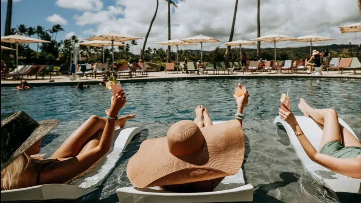 Three people enjoying a group getaway in Oahu, relaxing on poolside loungers, holding up drinks, and wearing large sun hats under a sunny sky. The pool is surrounded by umbrellas and palm trees, with other vacationers in the background.