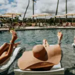 Three people enjoying a group getaway in Oahu, relaxing on poolside loungers, holding up drinks, and wearing large sun hats under a sunny sky. The pool is surrounded by umbrellas and palm trees, with other vacationers in the background.