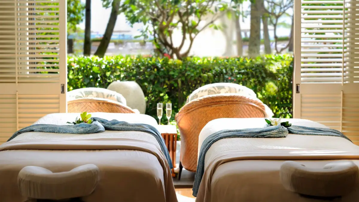 A serene spa room at the best spa hotel in Oahu features two massage tables draped in white linens, adorned with rolled towels and green leaves. Two wicker chairs and a small table with champagne glasses sit near large open windows, offering views of a lush garden outside.