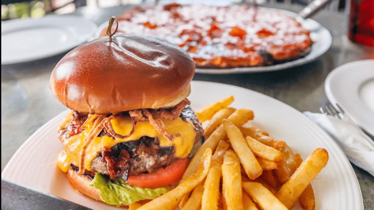 A juicy cheeseburger topped with crispy onions, lettuce, and tomato sits on a plate next to a serving of golden French fries. In the background, a pepperoni pizza is partially visible on another plate. Family-friendly restaurants on the South Shore ensure everyone enjoys these delicious meals.