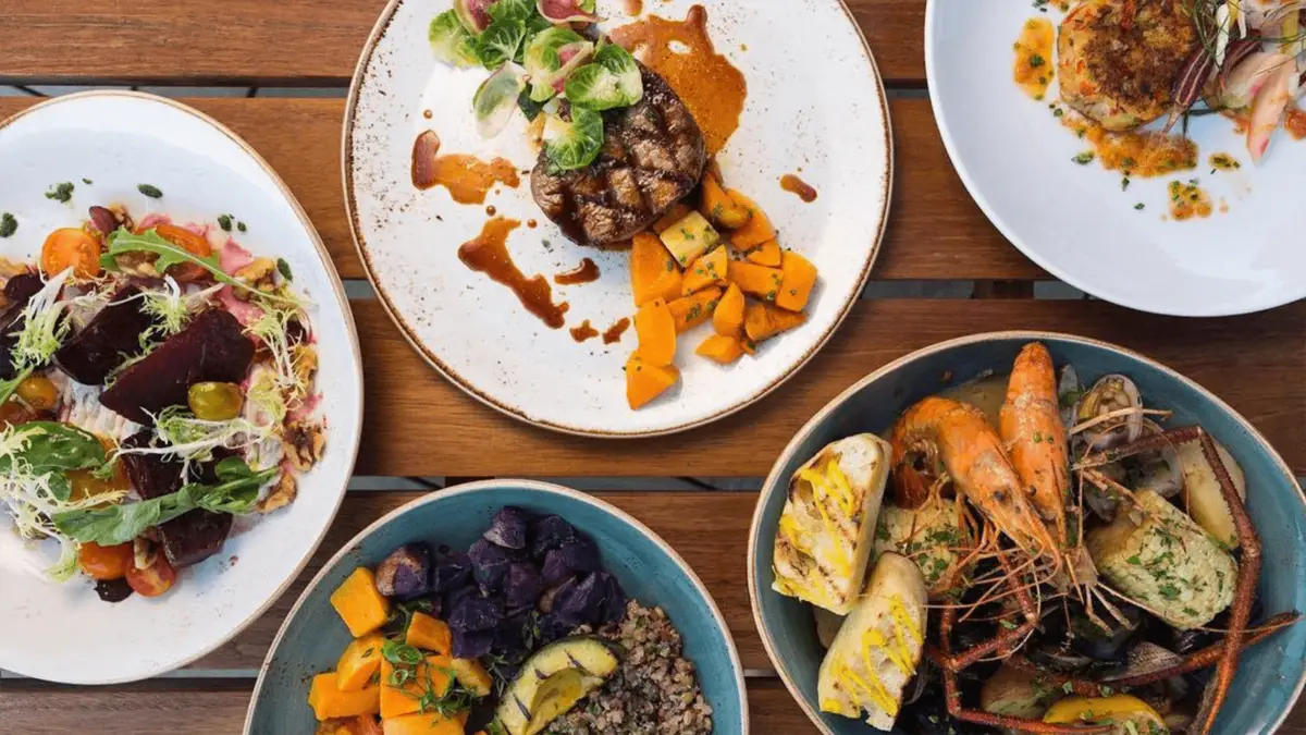 An overhead view of a wooden table set with five different plates of food: a plate with grilled steak and vegetables, a dish with seafood including shrimp, a bowl of mixed roasted vegetables, a salad with beets, greens, and tomatoes, and what appear to be the best gluten-free crabcakes on North Shore Oahu.