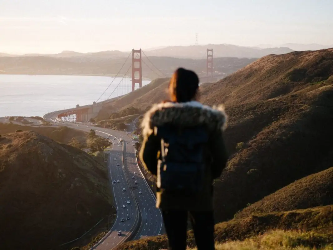 A person wearing a coat with a fur-lined hood and a backpack stands on a hill overlooking the Golden Gate Bridge at sunset. Below, cars travel along the winding road that leads to the bridge, surrounded by hilly, green terrain—truly one of the Bay Area's best things to do in March.