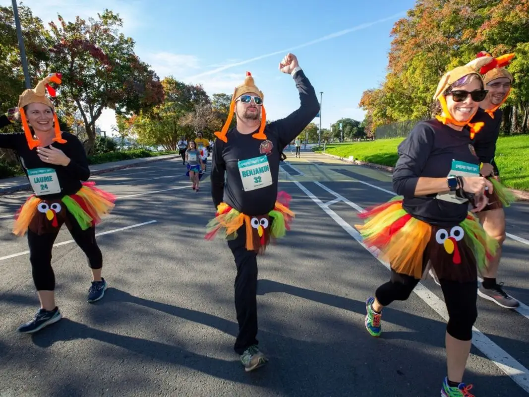Four people are participating in a fun run while wearing turkey-themed costumes, including colorful tutus and turkey hats. They look happy and are smiling as they run along a paved path with trees and greenery in the background—truly one of the best things to do East Bay November.