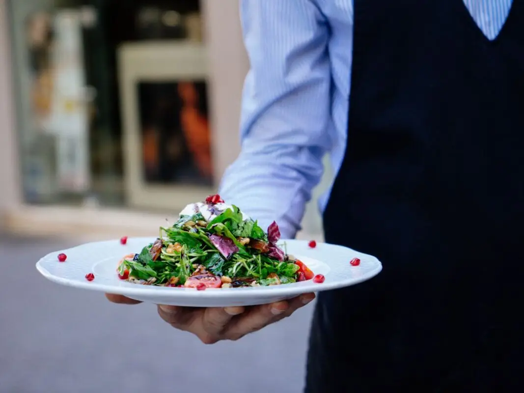 A person wearing a dark apron and light blue shirt holds a white plate with a fresh salad piled high with greens, pomegranate seeds, and various colorful vegetables. The background is slightly blurred, suggesting an outdoor dining setting—a perfect scene for one of the Best Things to do in the East Bay This March.