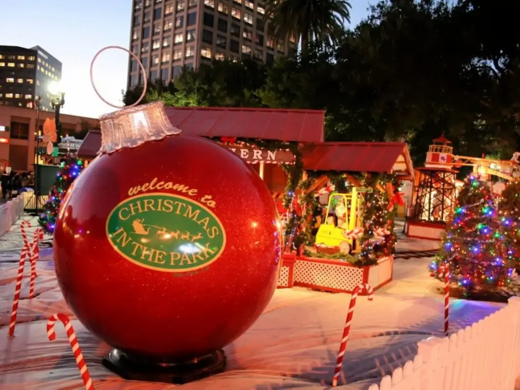 A large red Christmas ornament with the text "Welcome to Christmas in the Park" stands in front of festively decorated structures. Colorful lights adorn Christmas trees, and tall buildings are visible in the background, making it one of the best things to do in the South Bay this December.