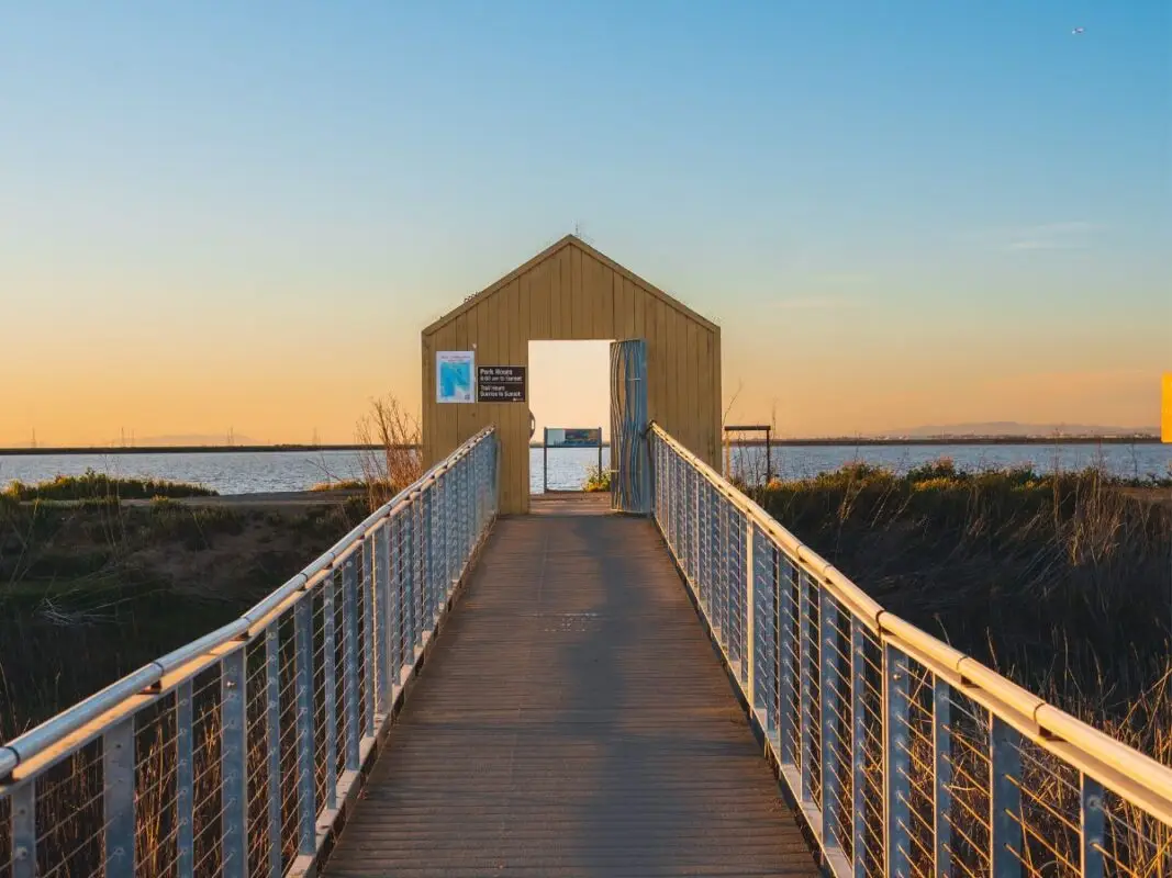 A paved path lined with metal railings leads to a beige, rectangular wooden structure overlooking a calm body of water. The sky is clear with a warm glow from the setting sun, casting a golden light on the scene. Signs are posted on the left side of the structure—one of the best things to do in South Bay this February.