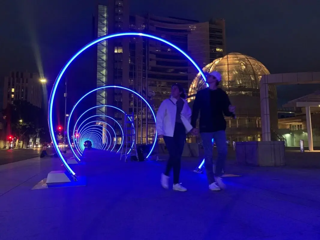 A man and woman walk hand in hand through an outdoor art installation featuring illuminated blue rings at night. The background includes a lit dome structure and tall buildings. This vibrant urban scene, one of the Best Things to do in South Bay in November, creates a captivating tunnel effect.