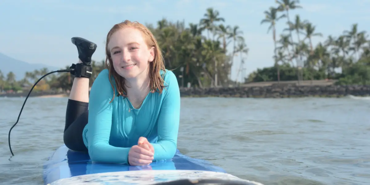 A person with wet hair and a blue rash guard is lying on a surfboard in the ocean, likely enjoying top surf lessons in Wailea. They are smiling at the camera, with one leg bent and the other resting on the board. The background features palm trees and a distant shore under a partly cloudy sky.