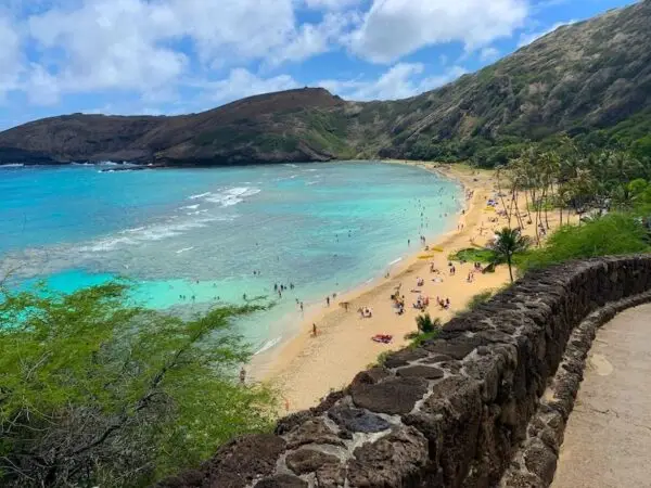 A stone wall runs along a path overlooking a scenic beach, one of the best things to do in Hanauma Bay. Clear turquoise waves gently lap against the sandy shore, where people are swimming and sunbathing. Verdant hills frame the background under a blue sky with scattered clouds.