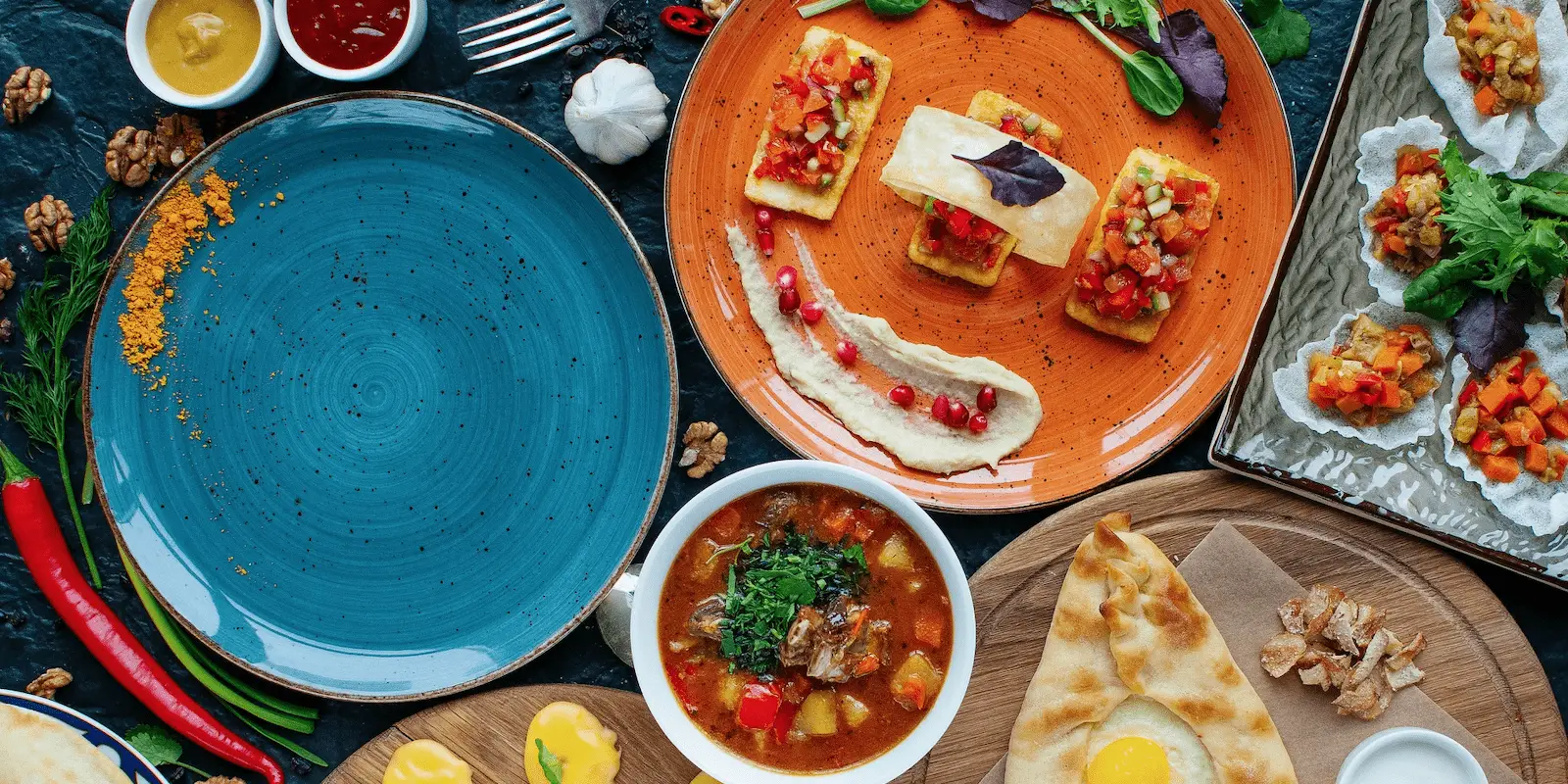 A table is set with a variety of colorful dishes: a blue plate with spice remnants, an orange plate with hors d'oeuvres and dip, a bowl of soup with herbs, flatbread with an egg, and a platter of assorted appetizers showcasing foods that define Hawaiian culture. Various condiments and garnishes are also present.