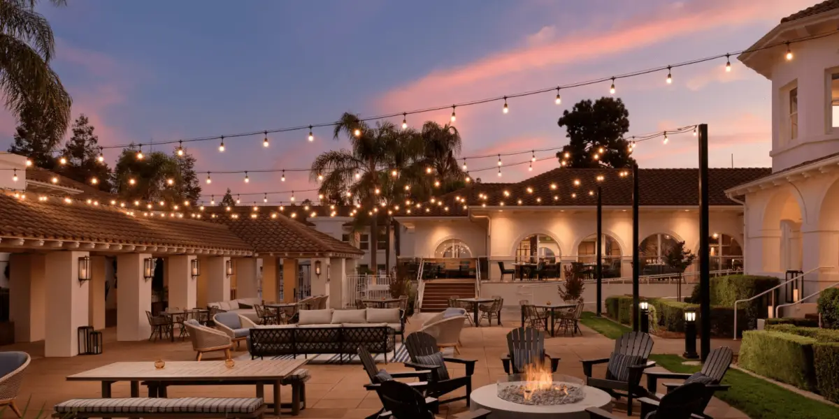 A picturesque outdoor patio at dusk featuring string lights overhead, cozy seating arrangements, a central fire pit, and palm trees scattered throughout. Reminiscent of the Best Spa and Wellness Hotels in the Bay Area, the setting exudes warmth and relaxation with a vibrant pink and purple sunset in the background.