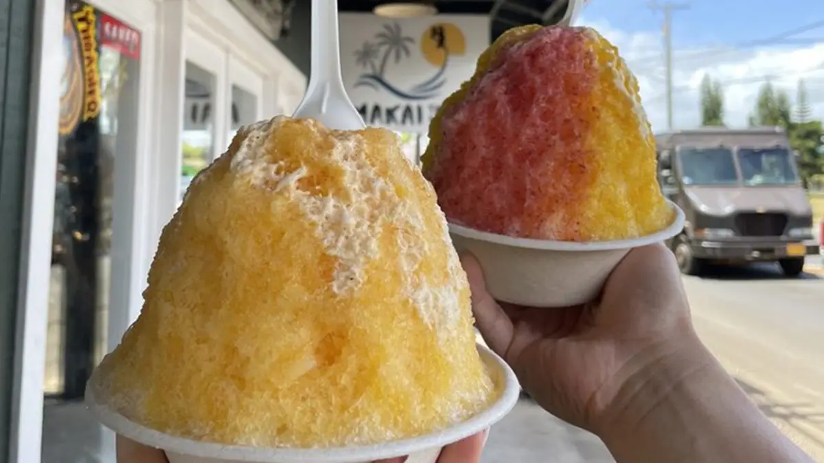 Two hands hold up cups of the best shave ice Kauai has to offer. The shaved ice on the left is yellow with a white spoon inserted in the top. The shaved ice on the right is a mix of yellow and red. A store sign and building are visible in the background.