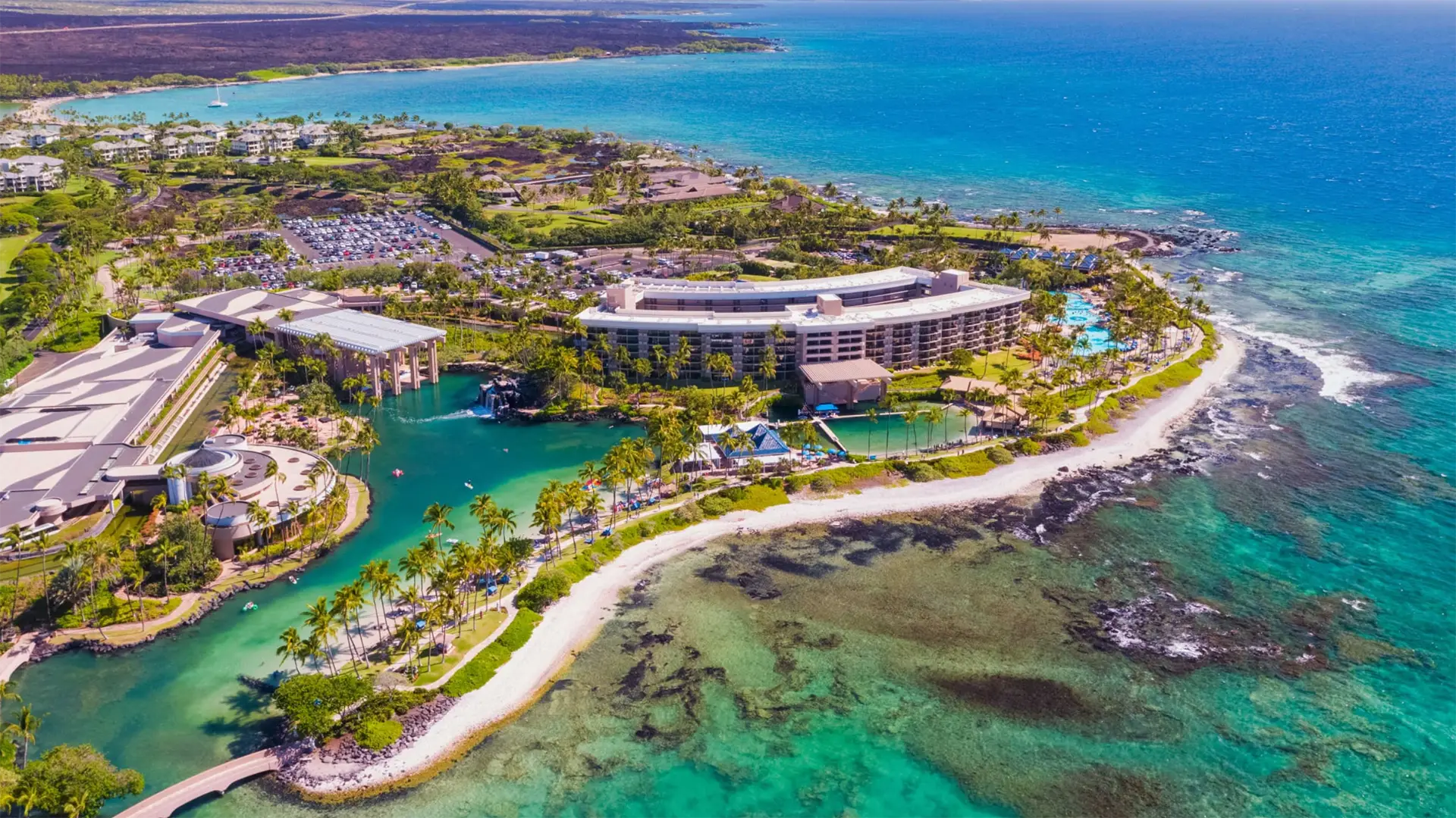 Aerial view of a coastal resort with multiple buildings set amidst lush greenery. Perfect for those seeking big island family hotels, the resort features various pools and is bordered by sandy beaches and clear turquoise waters. The surrounding area includes parking lots, scattered structures, and rocky shoreline.