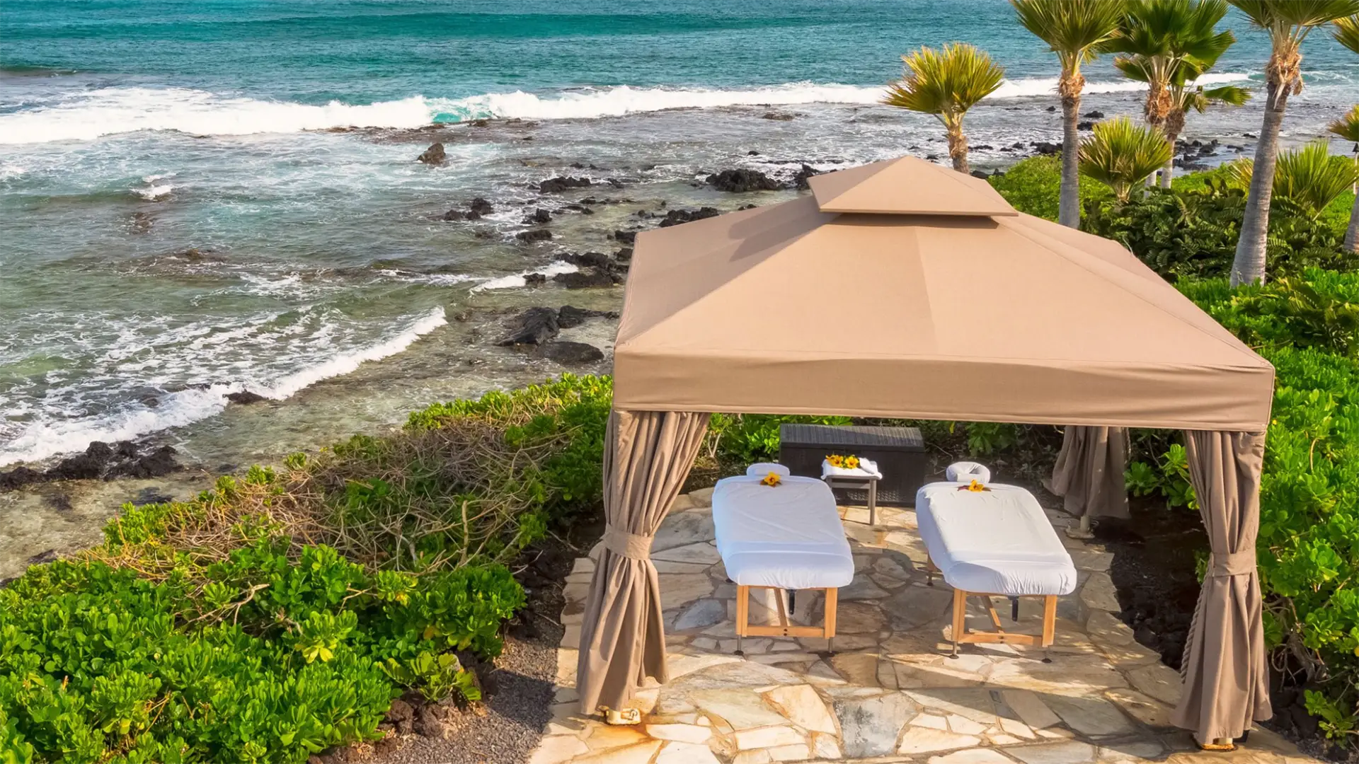 A luxurious beachside cabana at our hotel spa with a stone floor, housing two massage tables adorned with white sheets and small flower decorations. Surrounded by lush greenery, it overlooks a scenic ocean view with waves crashing against a rocky shoreline on the Big Island.