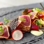 A gourmet dish of seared tuna slices artfully arranged on a slate plate awaits you at the best sunset dining spot on the Big Island. The tuna is garnished with thinly sliced radishes, green onions, and edible seaweed, accompanied by colorful vegetable cubes, a white sauce dollop, and a drizzle of black sauce.