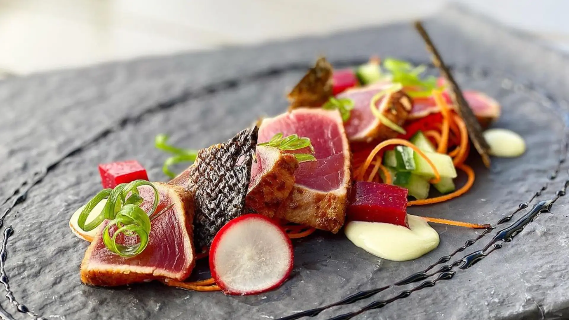 A gourmet dish of seared tuna slices artfully arranged on a slate plate awaits you at the best sunset dining spot on the Big Island. The tuna is garnished with thinly sliced radishes, green onions, and edible seaweed, accompanied by colorful vegetable cubes, a white sauce dollop, and a drizzle of black sauce.