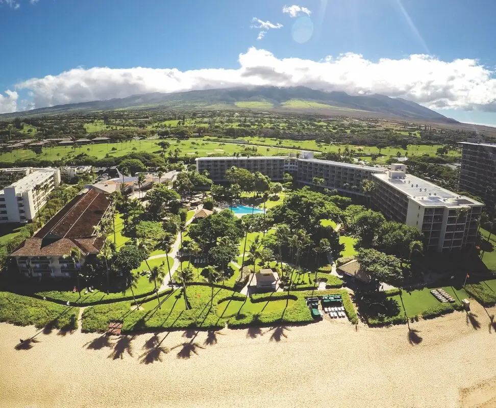 Aerial view of the Kaanapali Beach Hotel, a beachfront resort in Hawaii with multiple buildings surrounded by lush greenery. A large swimming pool is visible in the center. The resort is bordered by a sandy beach in the foreground with mountains and a partly cloudy sky in the background.
