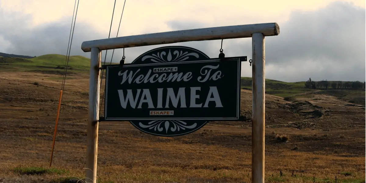 A wooden sign that reads "Welcome to Waimea" stands in a grassy field with hills in the background. The sky is cloudy, creating a serene and rustic atmosphere. Power lines are visible on the left side, and the sign includes the logo "ESCAPE," perfect for starting your food tour Waimea adventure.