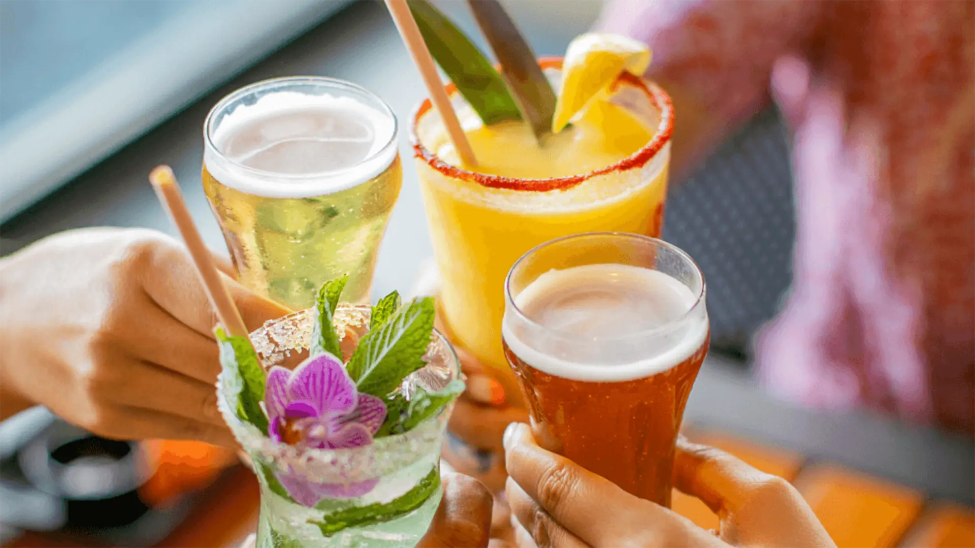 Four hands clink glasses in a celebratory toast, holding various drinks: a beer, a yellow cocktail garnished with lemon and leaf, a beer in a pilsner glass, and a minty cocktail with an orchid flower garnish. The background is blurred, evoking the lively atmosphere of the best happy hour Kauai has to offer.