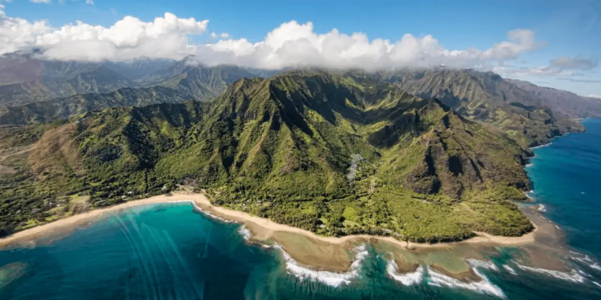 A scenic aerial view of a lush, green mountain range on the coast, with turquoise ocean waters washing up on sandy beaches. Rugged cliffs and dense vegetation are visible, while fluffy clouds hover over the peaks of the mountains in the background—truly the best island tour Oahu has to offer.